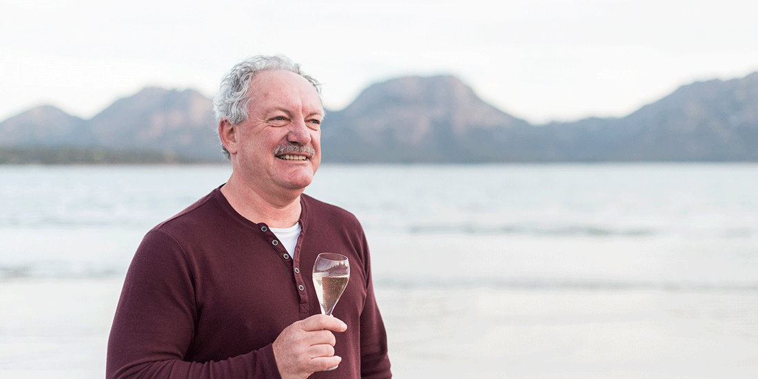 Man with grey hair and moustache standing in front of the ocean holding a glass of sparkling wine