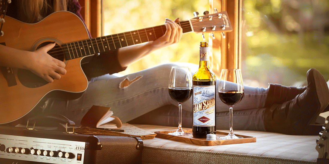 Close up of person playing acoustic guitar with a bottle and two glasses of red wine next to them