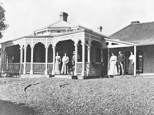 Black and white photograph of the exterior of a period home with six people standing outside