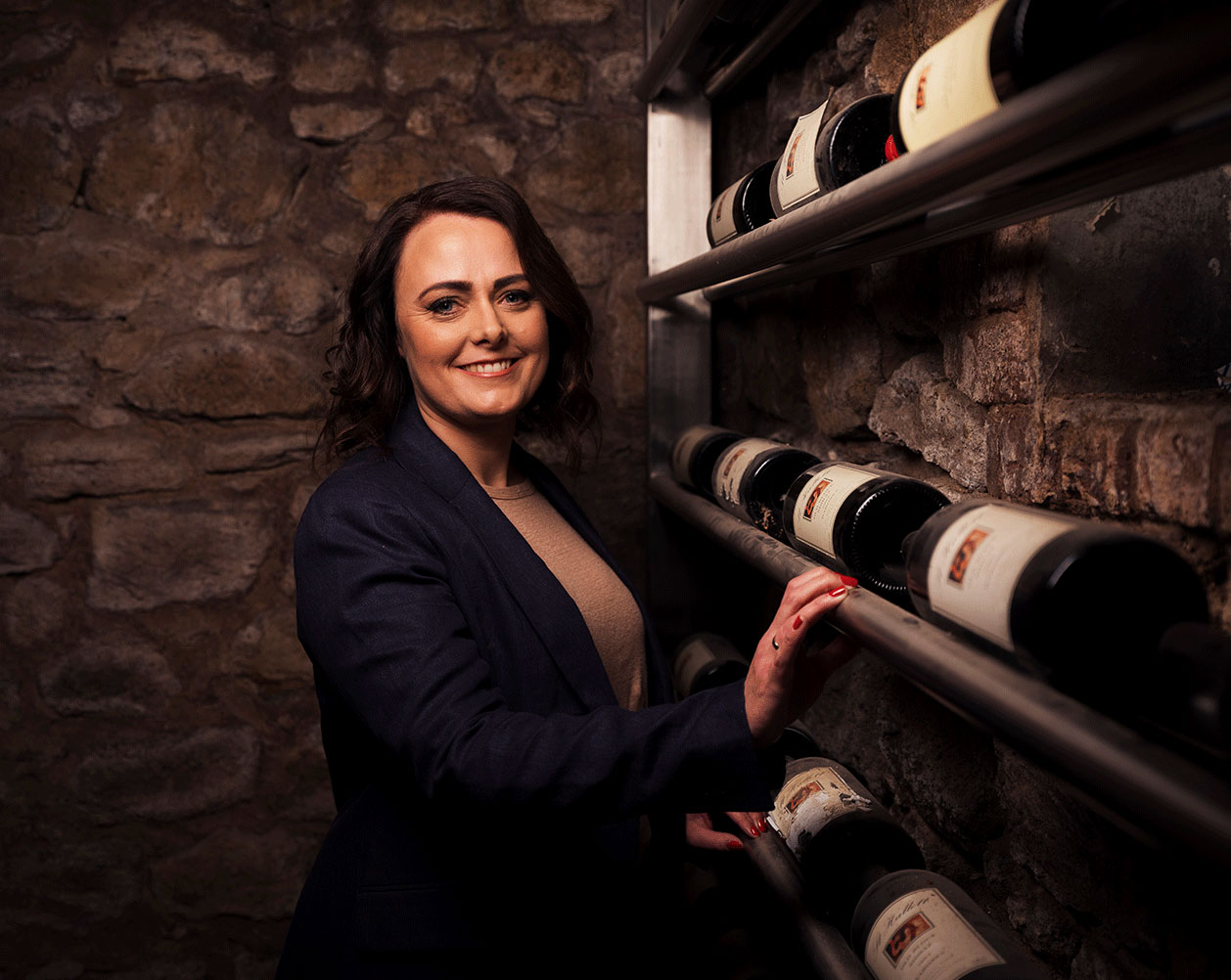 A smiling woman stands in a wine cellar in front of bottles