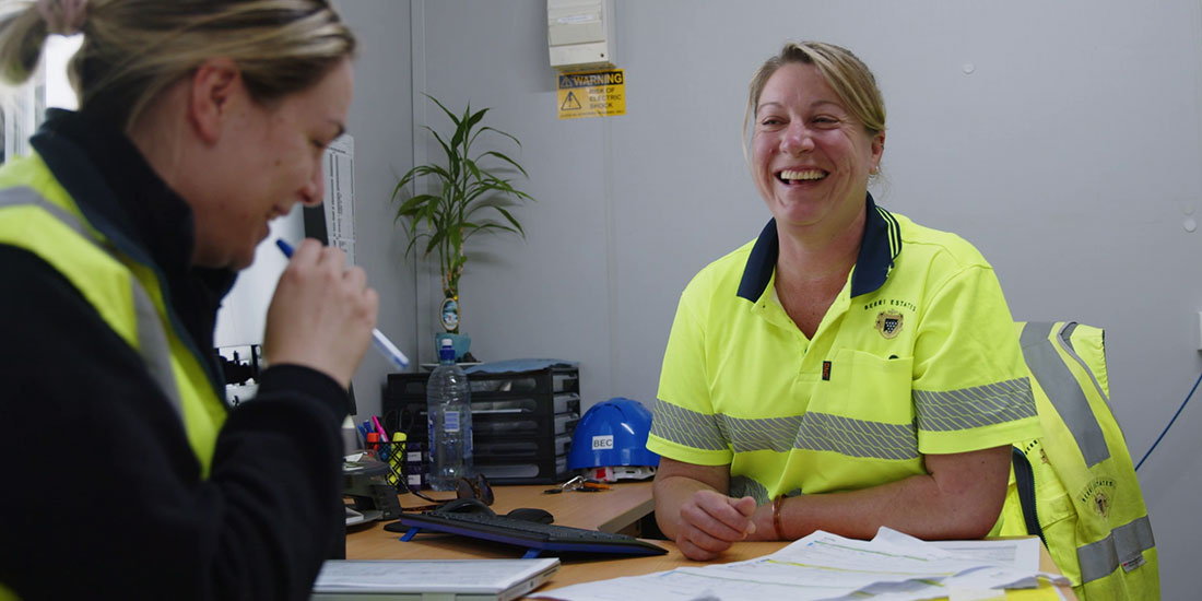 two women in high vis work wear sit at a table laughing