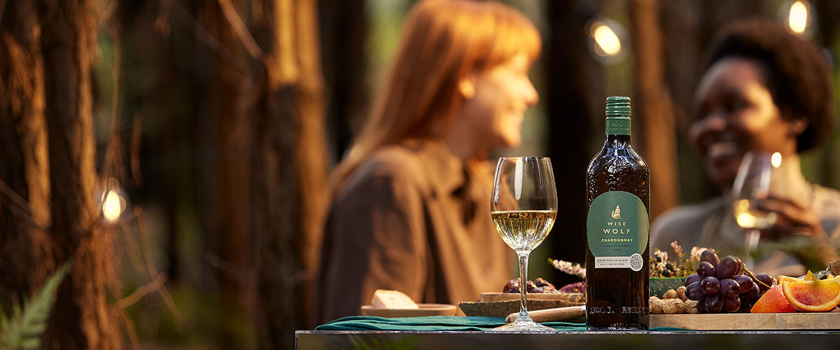 A Wise Wolf recycled glass bottle, with a glass of white wine on a mirror table in a forest of pine trees with 2 women talking in the background
